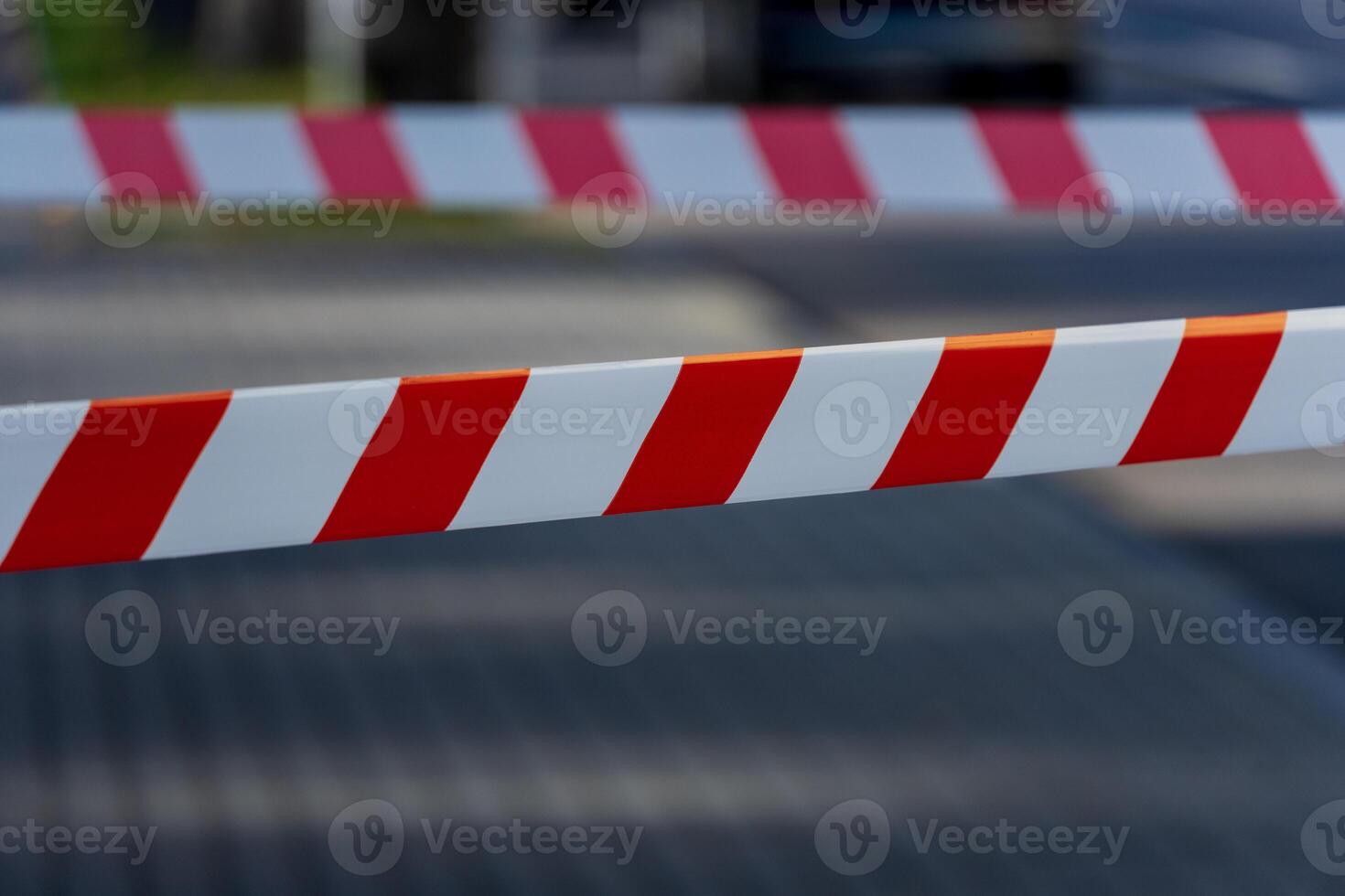Caution tape with bold red and white stripes stretches across a pathway, clearly marking a restricted area in a busy city setting. The sun casts shadows on the pavement photo