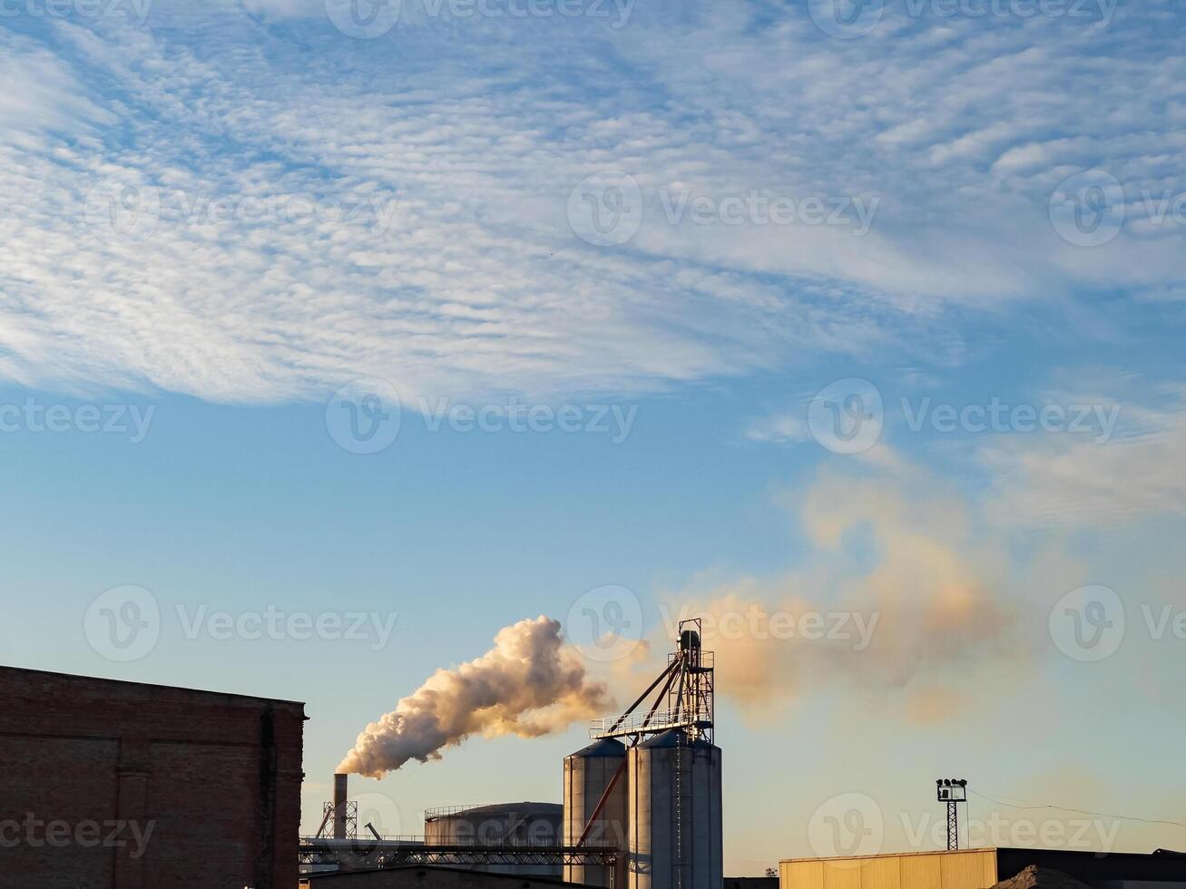 Thick smoke billows from smokestacks of a large factory, spreading out against a backdrop of cloudy blue sky, indicating active manufacturing processes photo