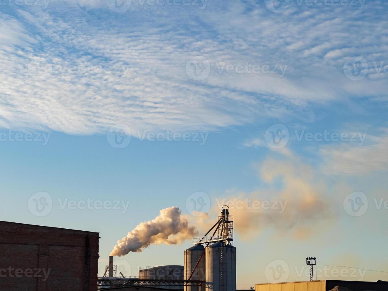 Thick smoke billows from tall smokestacks at a production facility under a cloudy sky. The scene showcases industrial activities, highlighting the environmental impact of manufacturing photo