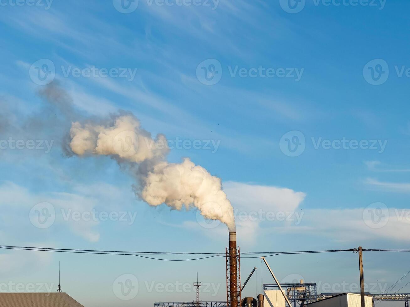 Factory operations produce large volumes of smoke from tall smokestacks, contributing to air pollution while buildings and machinery are visible beneath a clear blue sky photo