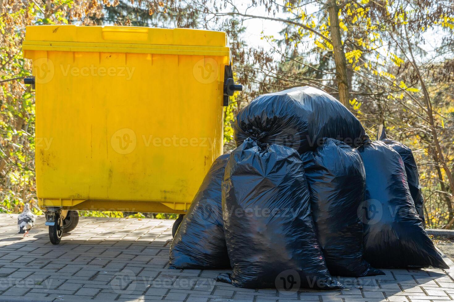 Black garbage bags are stacked near a vibrant yellow dumpster in a public area, highlighting issues of waste management and environmental conservation efforts in the community photo