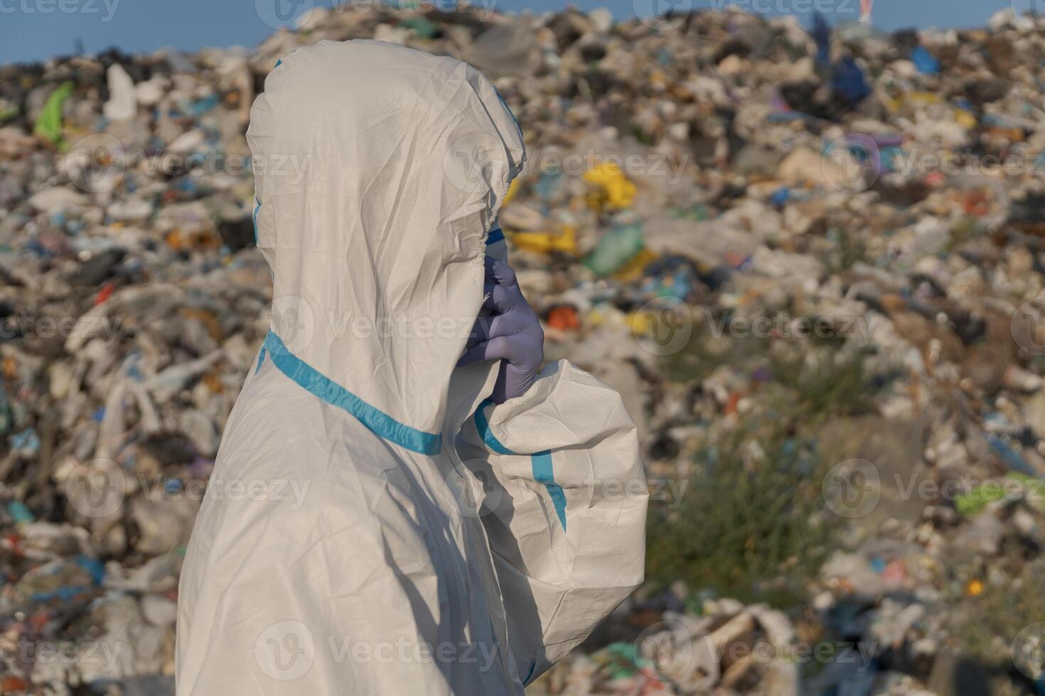 A worker in protective gear stands at a landfill, surveying a large area filled with trash and plastic waste under a clear sky. This highlights issues of pollution and ecology photo