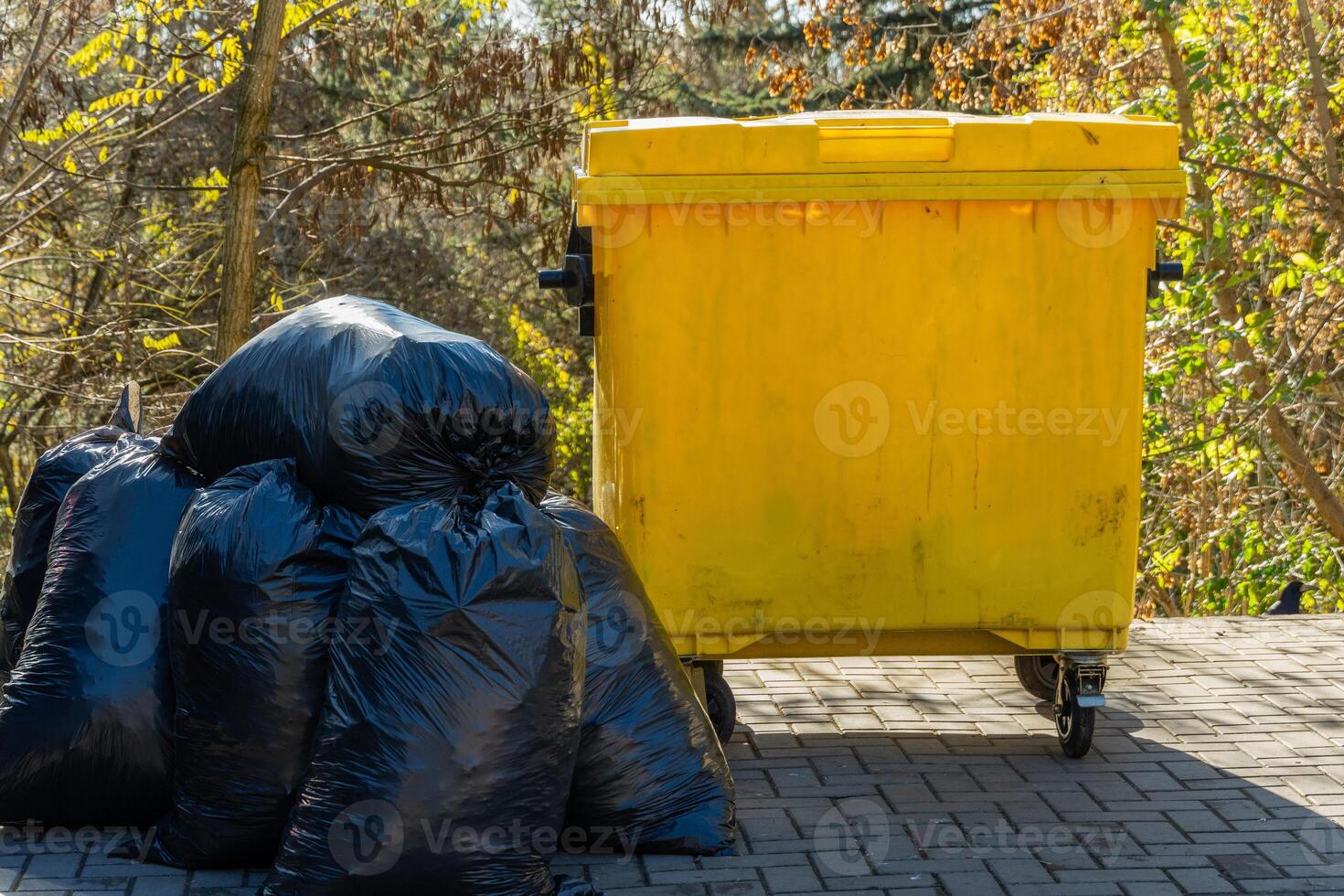 Black garbage bags are stacked next to a vibrant yellow dumpster in a wooded area, showcasing an important aspect of waste management and environmental conservation photo