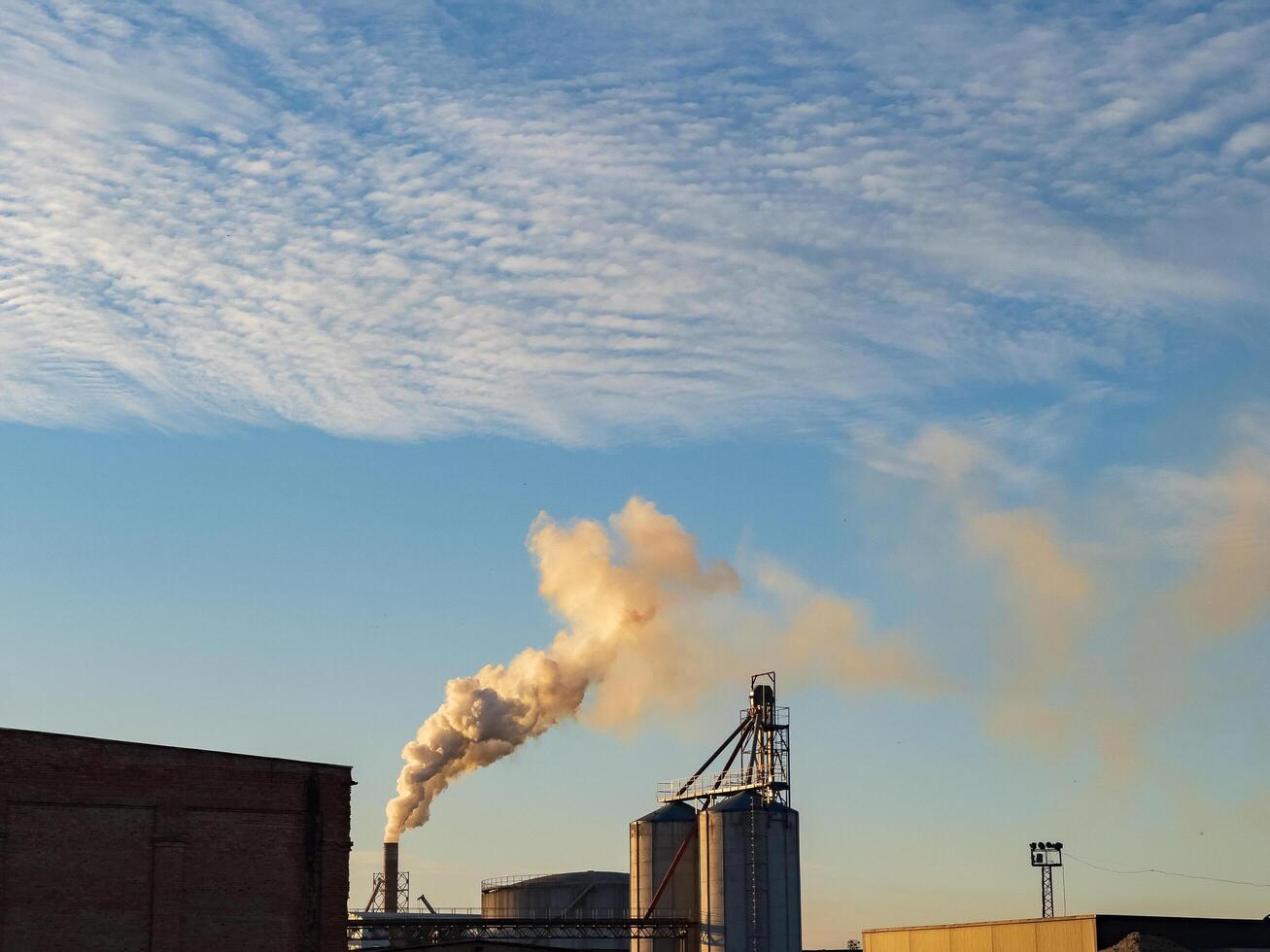 In a sprawling industrial area, smokestacks release plumes of smoke against a backdrop of fluffy clouds. The warm hues of sunset cast a golden light on the structures photo
