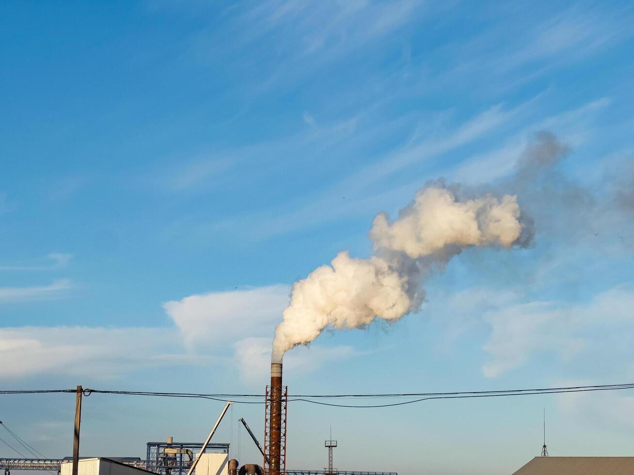 In the tranquil early morning, a plume of white smoke escapes from a tall industrial chimney, contrasting with the vivid blue sky. The peaceful atmosphere hints at activity beginning anew photo