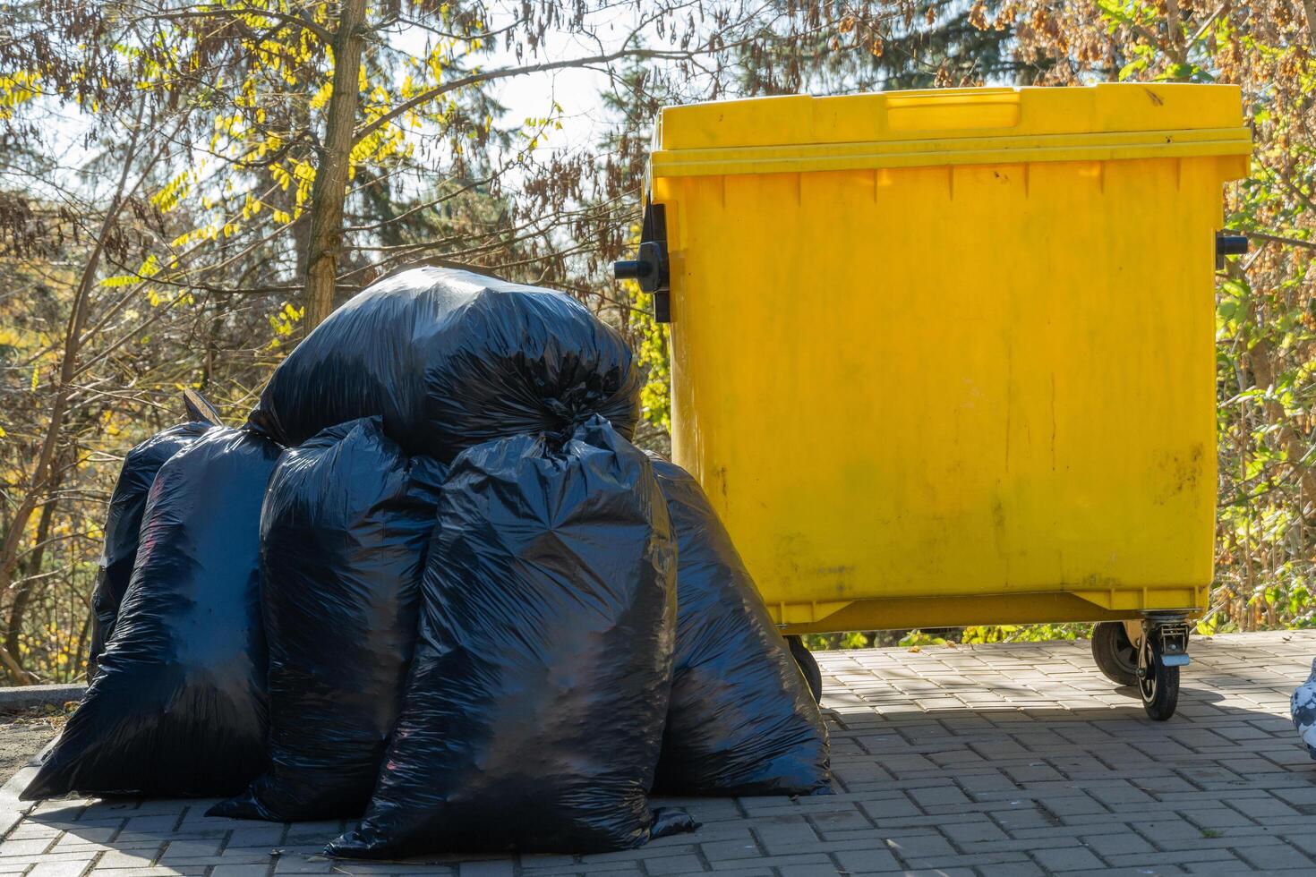 Black garbage bags overflow near a bright yellow trash bin, set against a backdrop of trees and sunlight filtering through leaves, highlighting urban waste management efforts photo