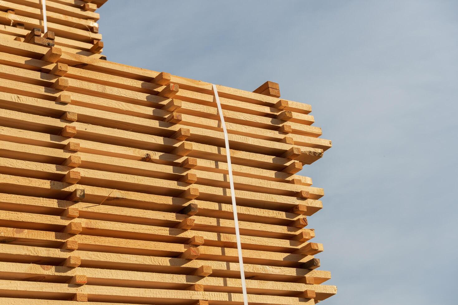 A neatly organized pile of wooden planks rises against the backdrop of a bright blue sky. The timber is tightly bundled, showcasing rich textures and natural colors under the sun photo