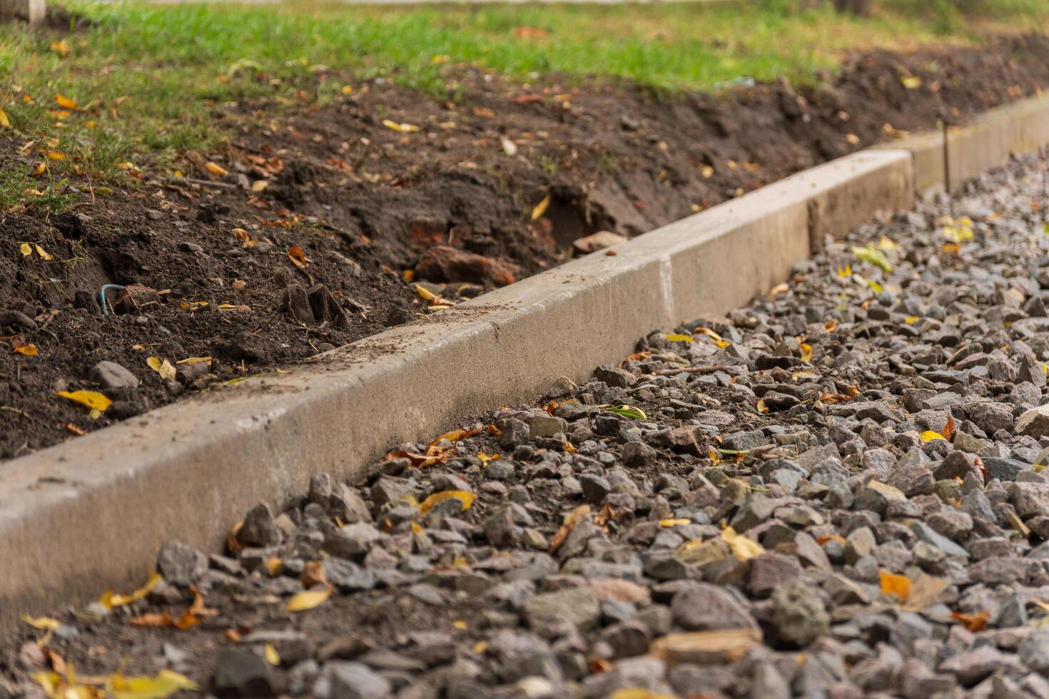 A newly installed concrete curb lines a gravel pathway, with earthy tones blending into the surrounding greenery. Fallen leaves add an autumn vibe to the tranquil atmosphere photo