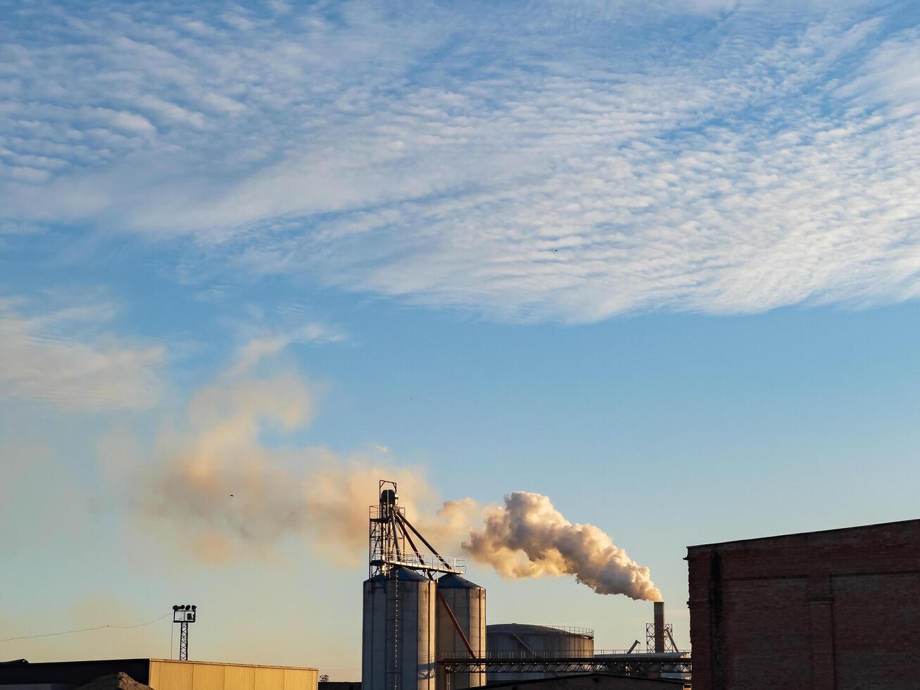 Industrial silos stand tall as smoke rises into the crisp evening air, contrasting with the bright colors of the setting sun and the textured sky above photo