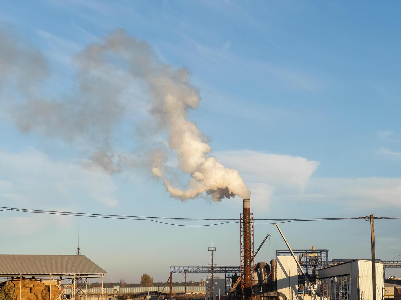A factory actively emits white smoke from its chimney, contrasting with the vibrant blue sky above. The surrounding setting showcases industrial infrastructure and a serene environment photo