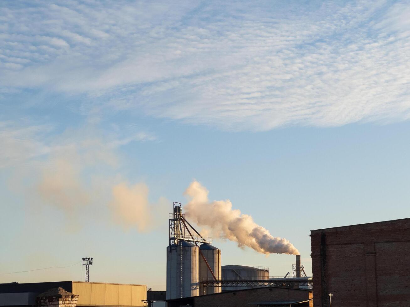 Against a backdrop of a stunning blue sky, towering smokestacks release puffs of smoke as the sun sets, casting a warm glow over the industrial landscape and evoking a sense of transformation photo