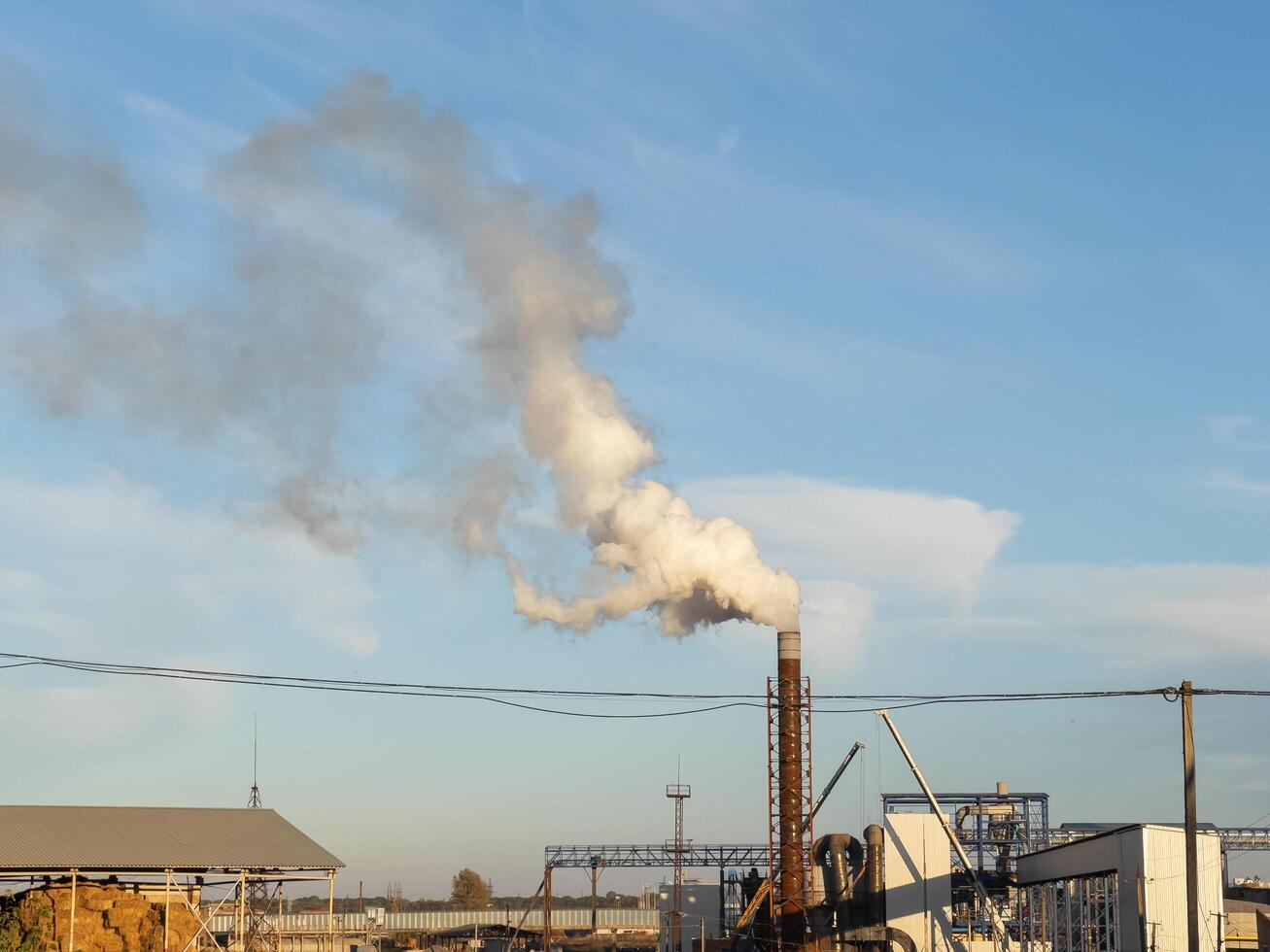 A plume of smoke rises from a tall chimney at a manufacturing plant, contrasting sharply with the clear blue sky. The sun's warm rays illuminate the scene during late afternoon photo