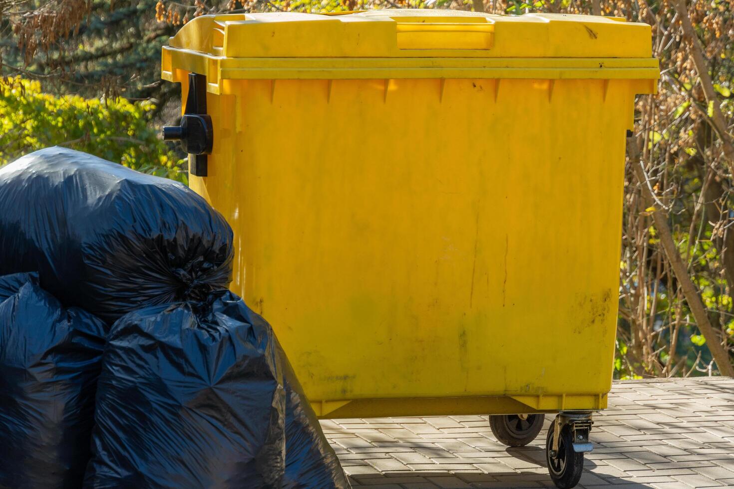A vibrant yellow garbage can stands in a tranquil urban park, accompanied by neatly stacked black garbage bags, enjoying the warmth of a sunny afternoon photo