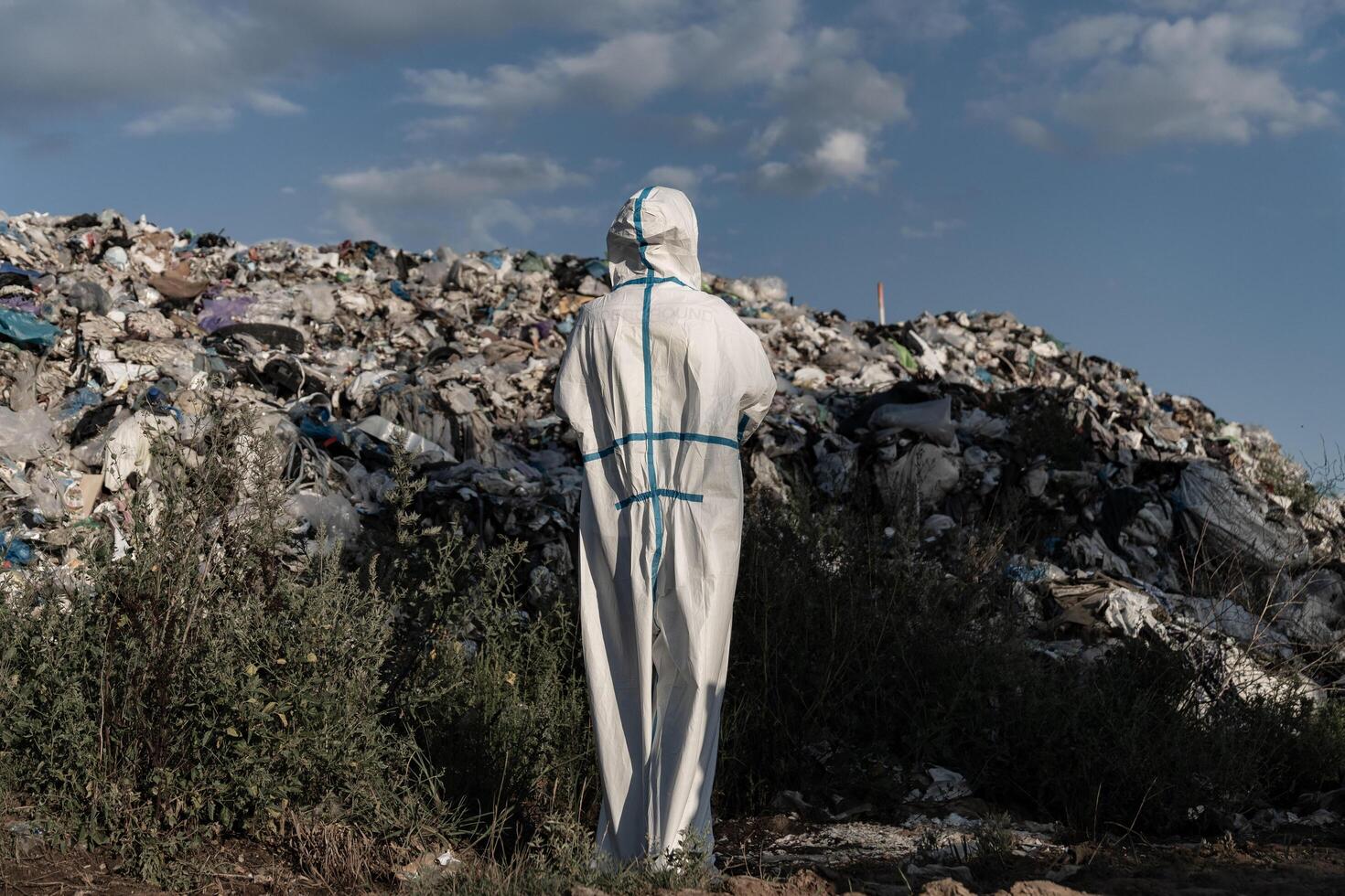 Wearing a protective suit, a worker stands before a towering mountain of waste, surveying the landfill's expansive waste and debris under a blue sky photo