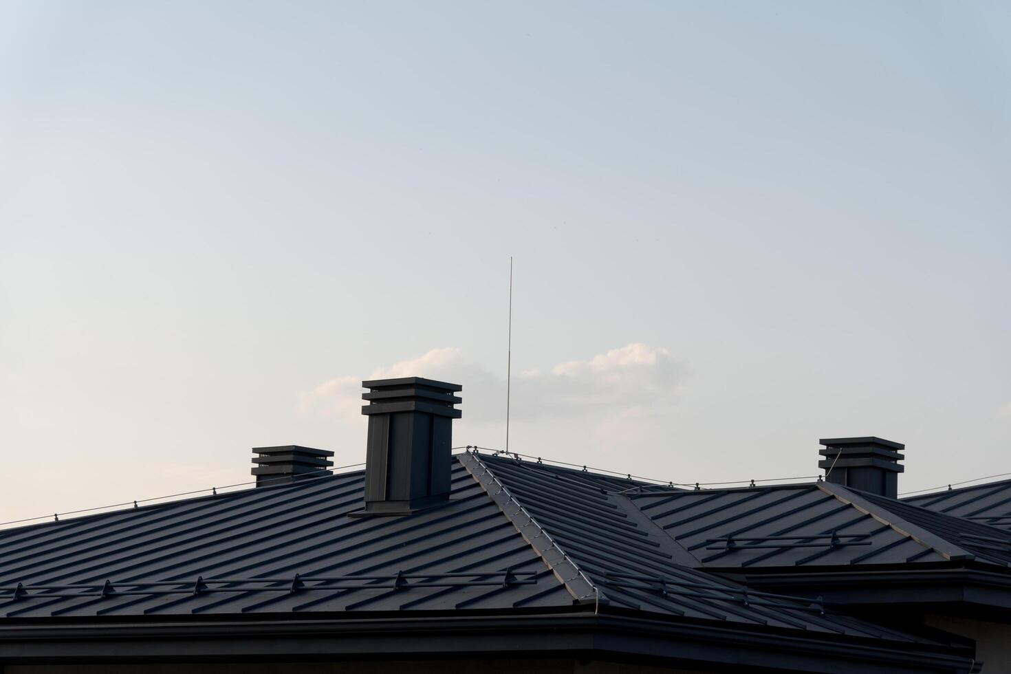As the sun sets, a modern home's distinctive metal tile roof reflects soft hues of the sky. Tall chimneys add character while a delicate antenna reaches for the clouds, evoking tranquility photo
