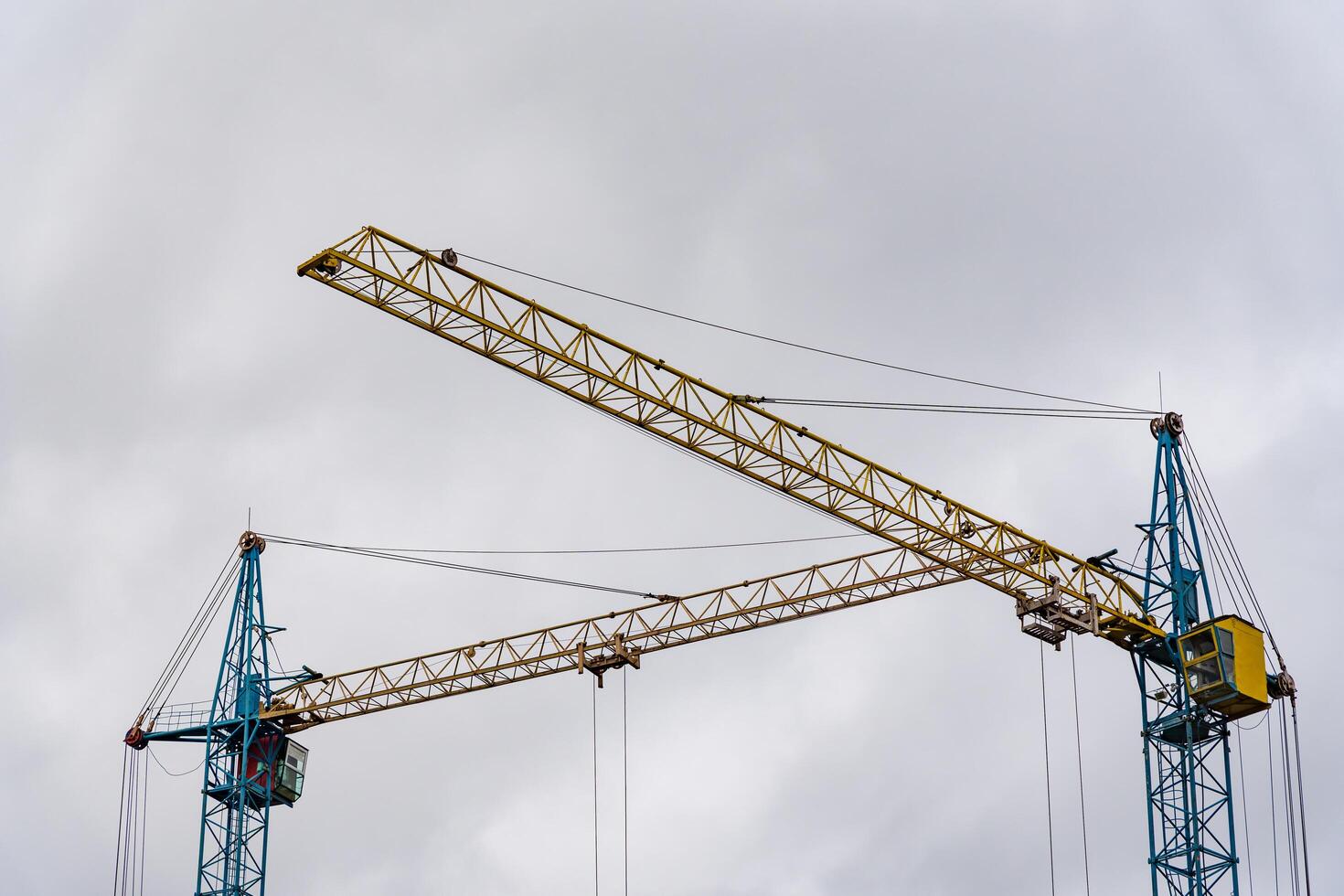 Two large construction cranes are actively positioned at a construction site, lifting materials as workers prepare the framework for a new house. The sky above is overcast photo