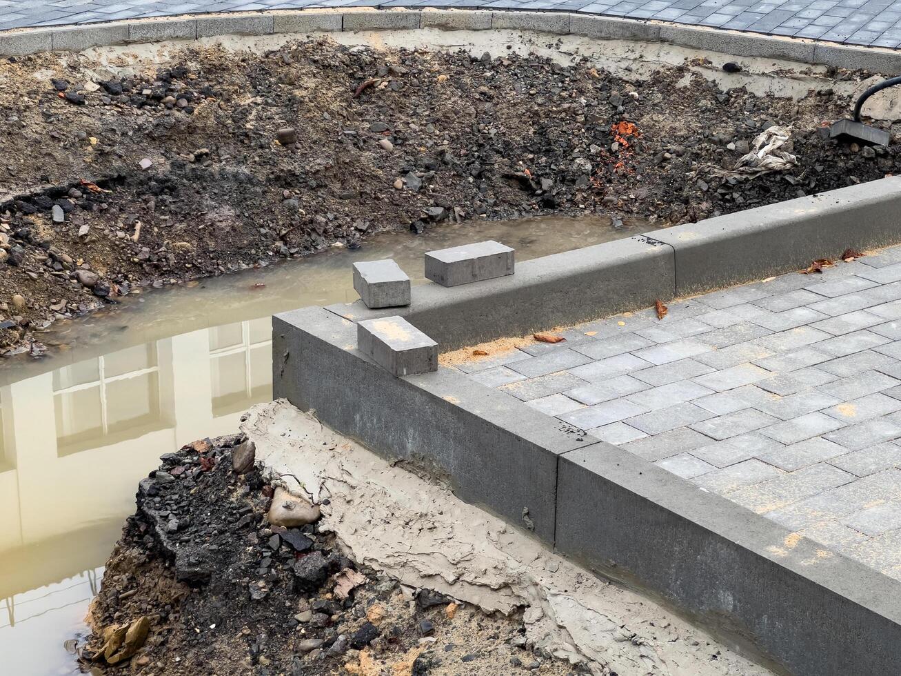An unfinished brick pavement stretches alongside a sidewalk, featuring a trench filled with water. Loose soil and uninstalled pavers indicate ongoing construction activity photo