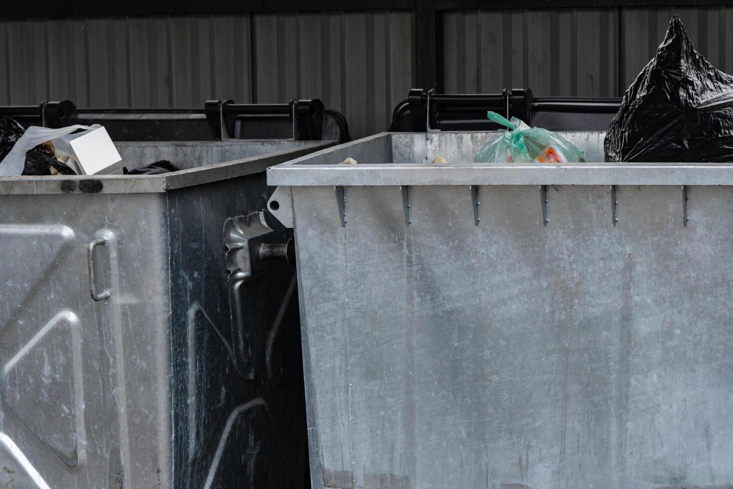 Two metal garbage containers filled with various types of rubbish are situated in a covered public space, collecting waste during daylight hours. The area shows signs of urban disarray photo
