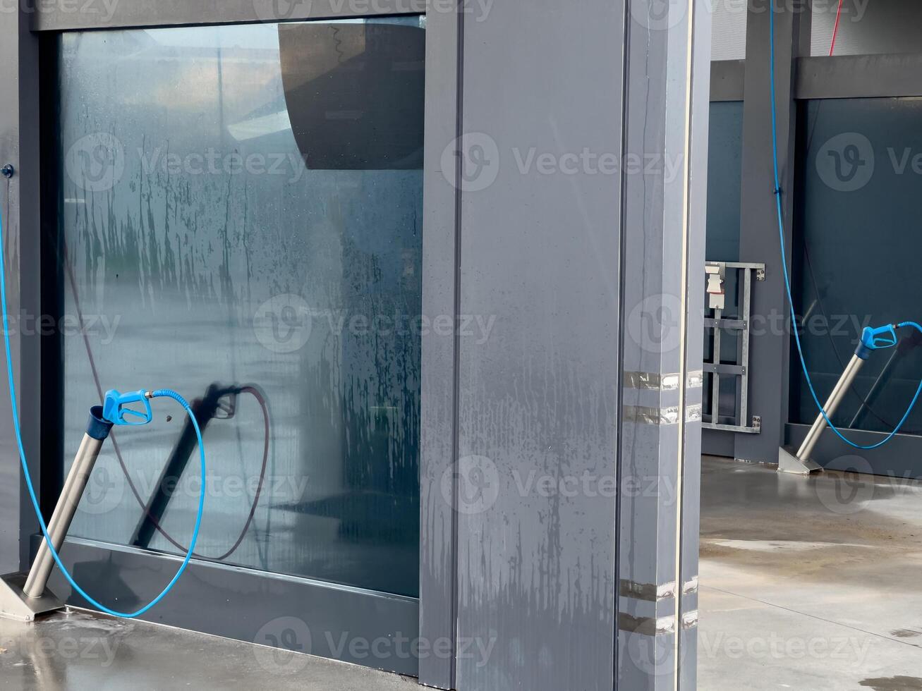 A view of a car wash station. The station is made of grey metal. There are two long blue hoses attached to high-pressure sprayers in front of a clear glass door photo