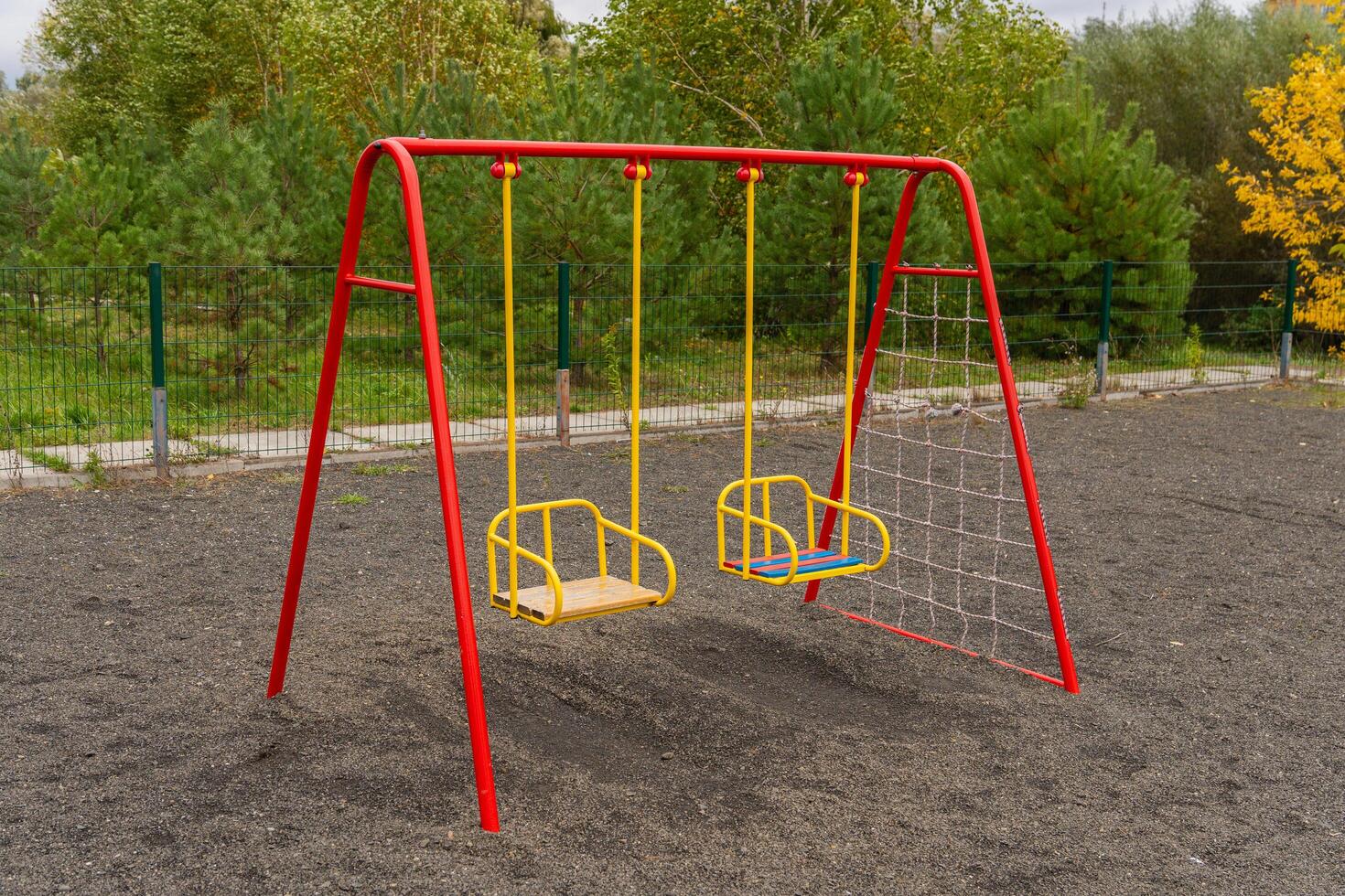 Two brightly colored swings sit in a park with soft ground cover, surrounded by lush greenery, perfect for children to play and enjoy a sunny day outdoors photo