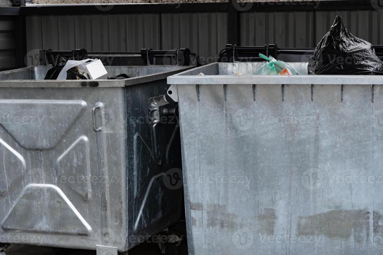 Two metallic garbage containers are positioned in a municipal area, overflowing with various types of waste, including bags and discarded items during daylight photo