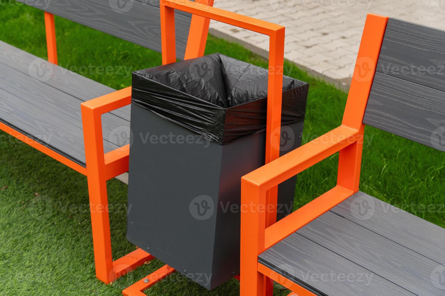 A black garbage can with a black plastic liner is mounted to an orange bench. The bench is made of metal with a gray wooden top. The bench is situated on a green lawn photo