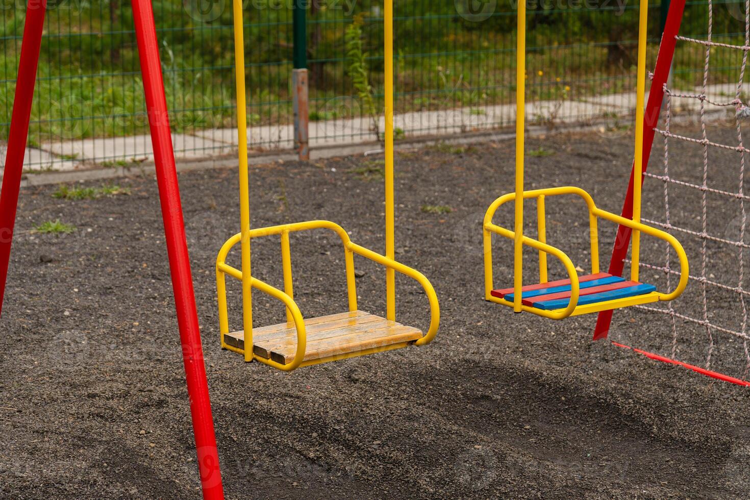 Two yellow swings with red supports stand still on a playground. The ground is made of gravel. There is a net fence behind the swings and green trees in the background photo
