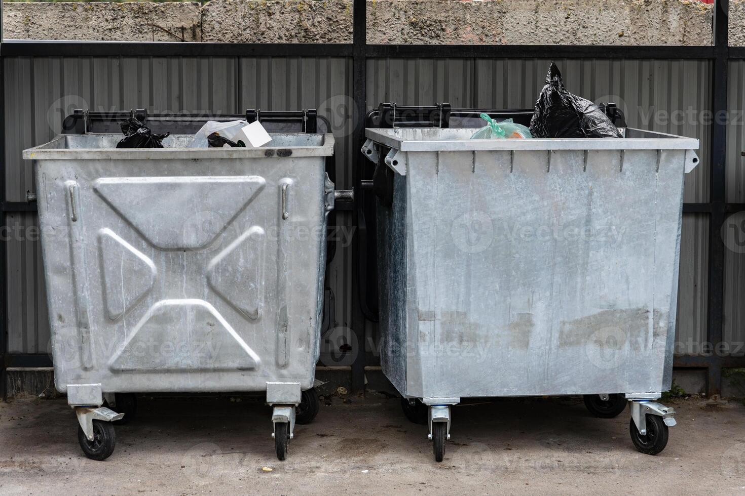 Two silver garbage containers are positioned on wheels, filled with black plastic bags and assorted waste, located in a busy urban alley under clear daylight photo