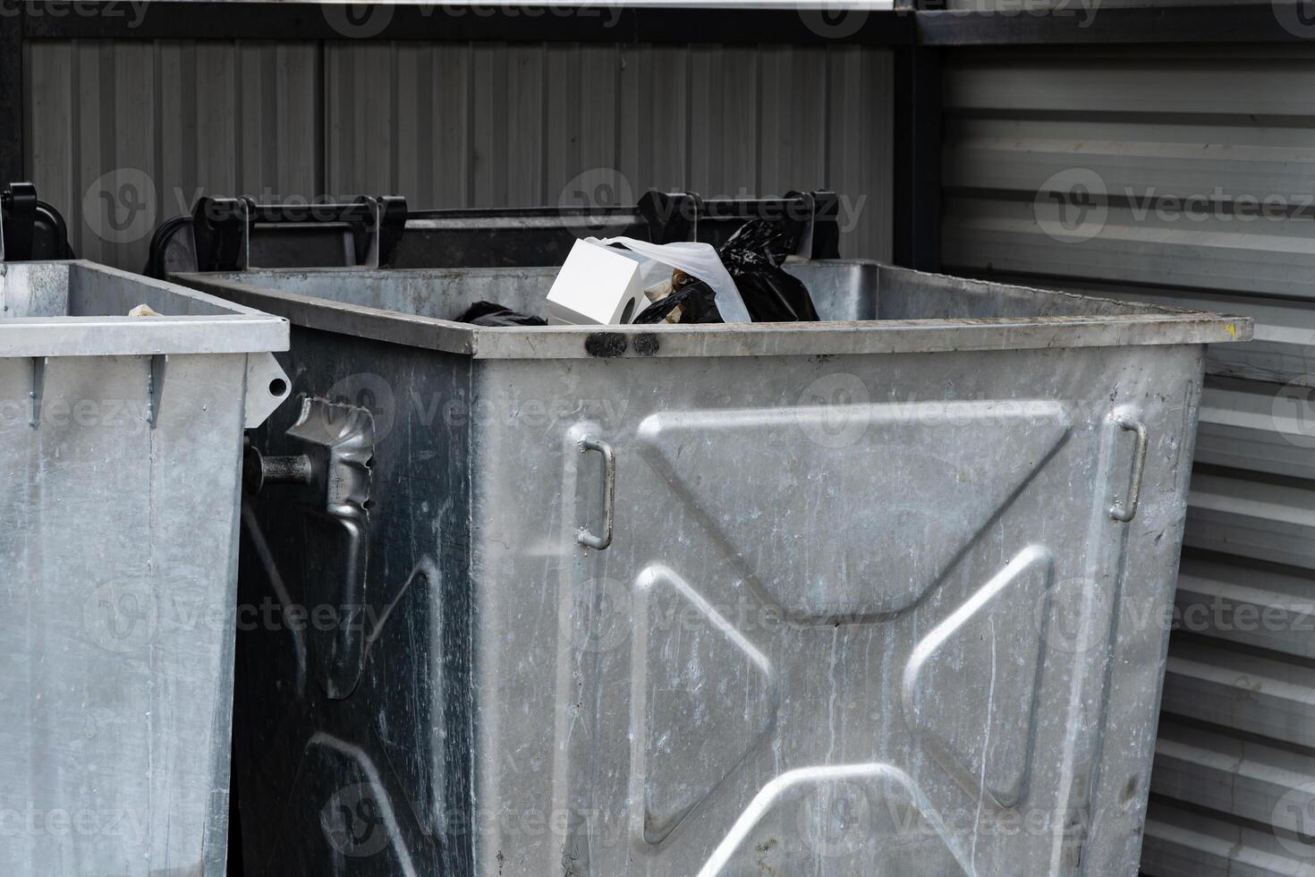 Two metallic garbage containers are positioned side by side, overflowing with various discarded items, captured during daylight in a city environment photo