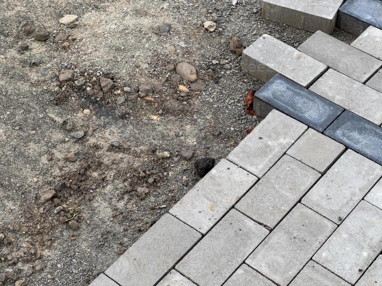 A section of pavement is being constructed, showcasing gray interlocking bricks. Nearby, an unfinished area of gravel and loose stones is visible, indicating progress in development photo