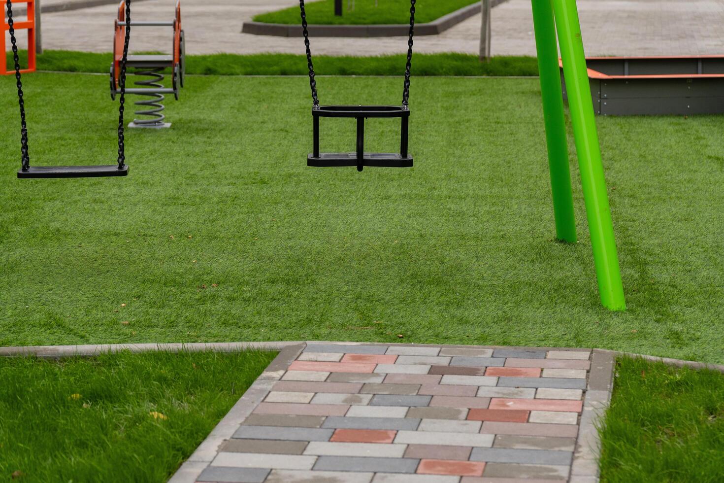 The playground features vibrant swings set on a soft green surface, surrounded by pathways, ready for children to explore, play, and have fun in the sun photo