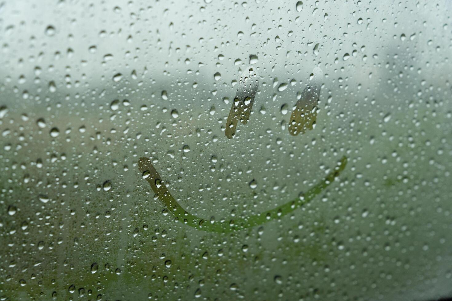 A simple smiley face is drawn on a foggy car window, surrounded by droplets of rain. The artwork brightens the dreary atmosphere on a rainy day photo