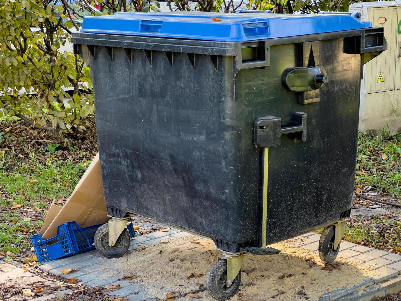 A black trash can with a blue lid rests on a paved city street. The can has four wheels and is made of plastic photo