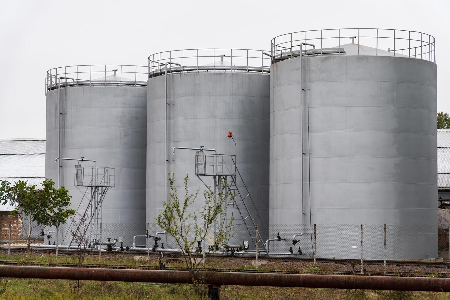 Three large, gray cylindrical storage tanks are positioned next to one another, equipped with metal ladders and catwalks, indicating an industrial environment photo