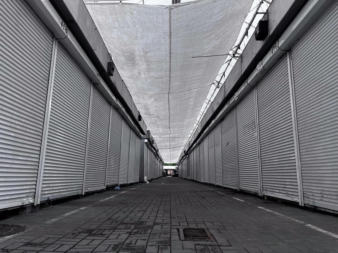 The narrow passageway of a once-busy market stands silent, lined with shuttered stalls. The overhead canopy casts shadows on the vacant ground, hinting at a forgotten vibrancy photo