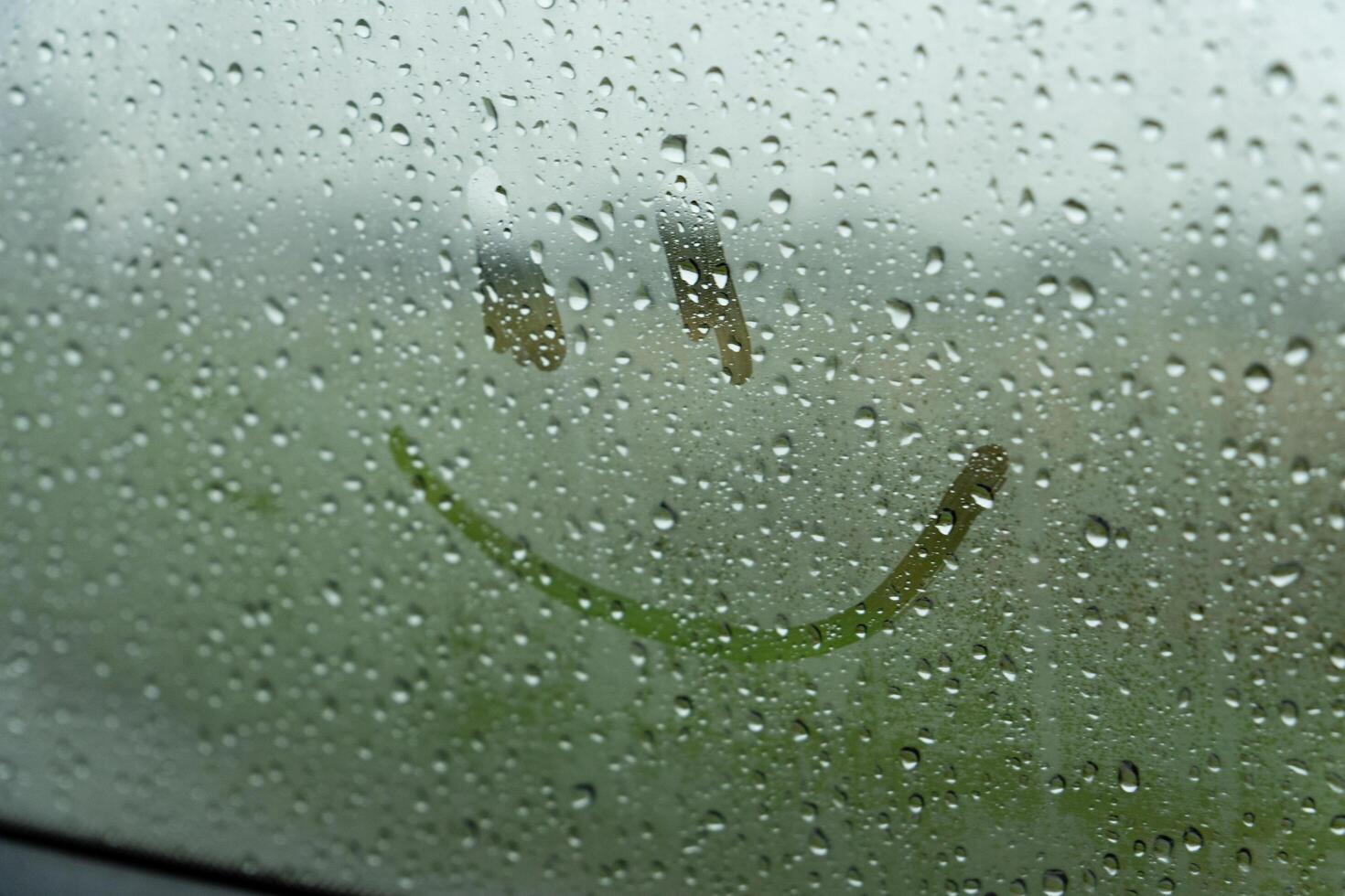 A single, simple smiley face is drawn on a car window. The window is covered in raindrops, obscuring the view outside. The smiley face is visible through the water droplets photo