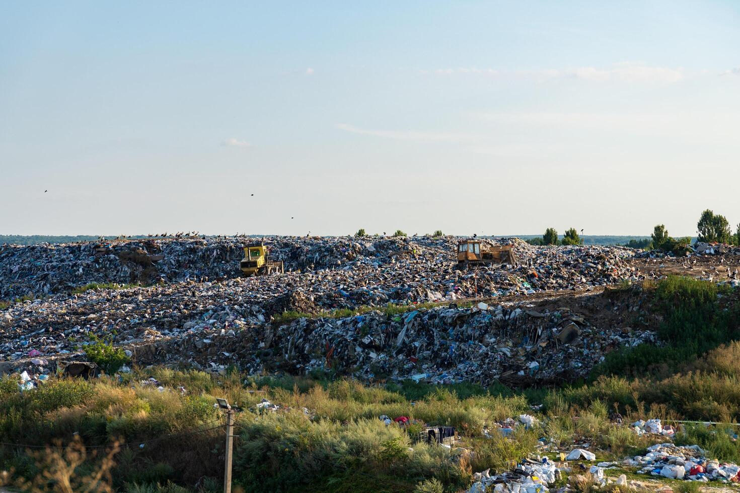 Heavy machinery works on a sprawling landfill surrounded by greenery, showcasing the scale of waste management in this rural environment during sunny weather photo
