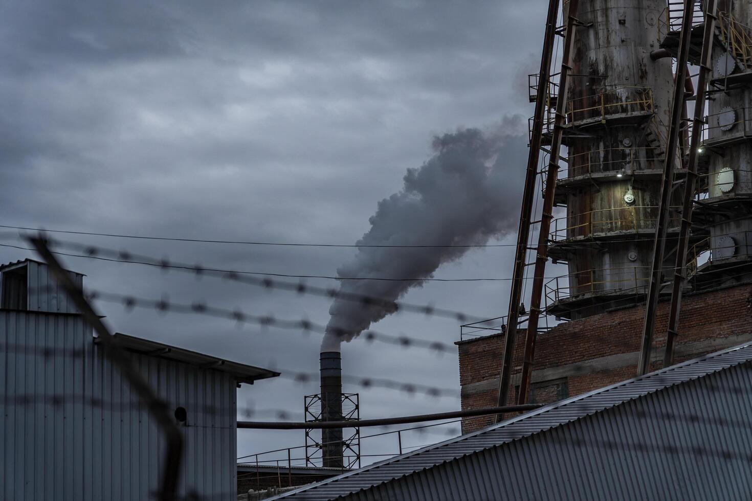 A large smokestack releases thick smoke into the air, surrounded by industrial buildings. The overcast sky adds a somber mood to the late afternoon scene photo