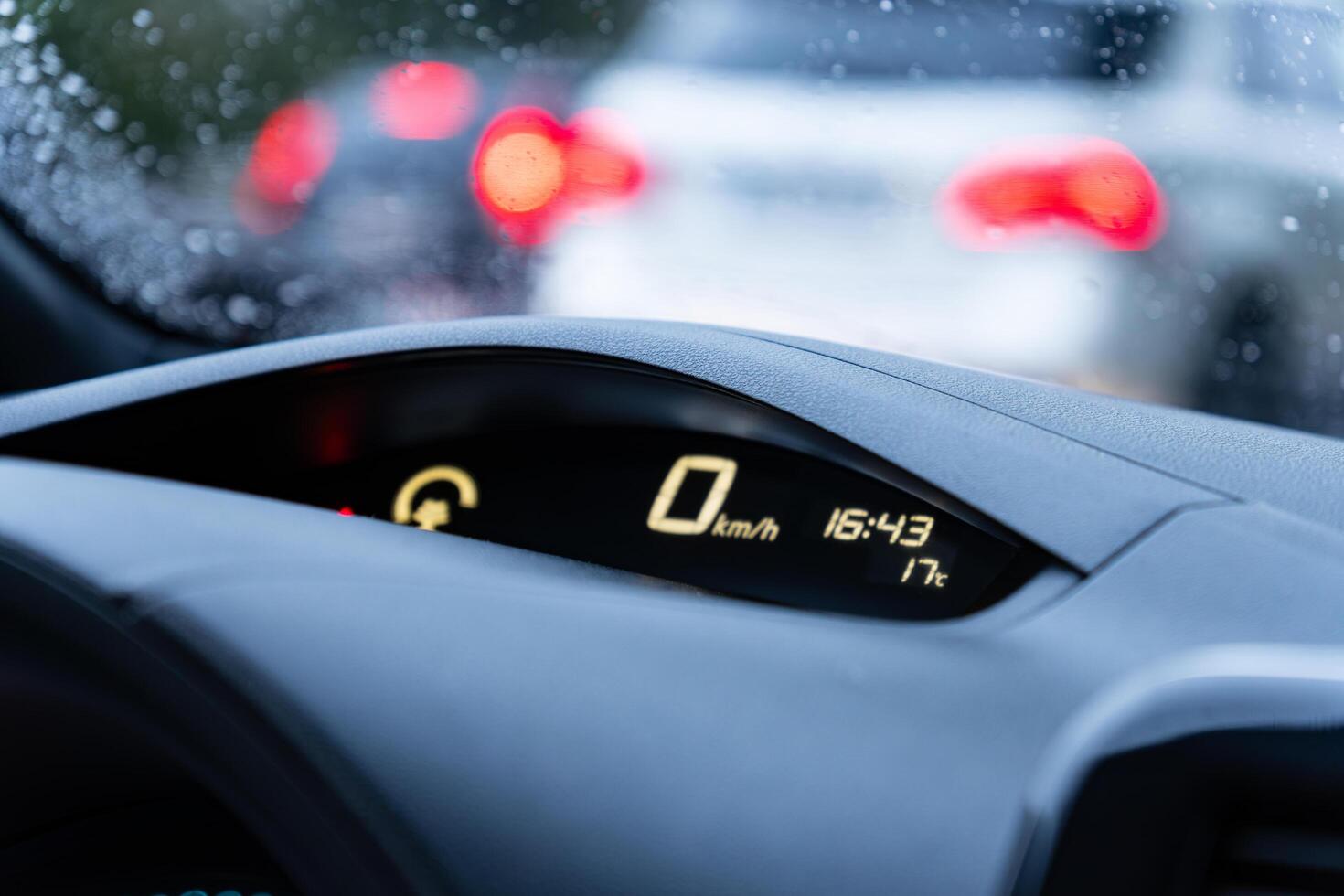 The dashboard shows zero kilometers per hour, indicating stopped traffic during rain, while cars are blurred in the background, creating a sense of delay photo