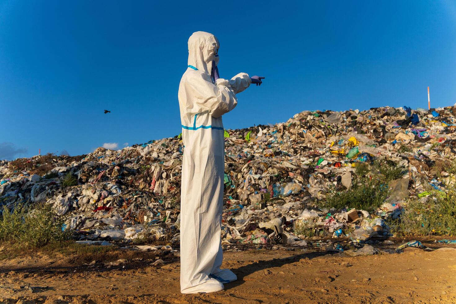 A person dressed in a protective suit stands at a landfill, looking out over heaps of waste, surrounded by blue skies and scattered debris, emphasizing environmental concerns photo