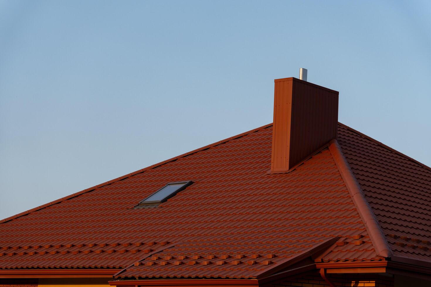 The red-tiled roof of a house features a skylight and a chimney, set against the backdrop of a clear blue sky, highlighting the architecture and design photo