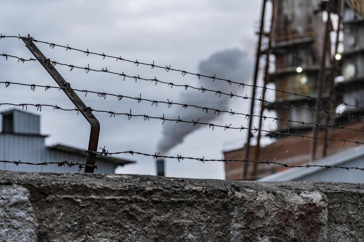 A barbed wire fence stands in the foreground, with a factory in the background exhaling smoke into the gray sky, reflecting an industrial environment photo