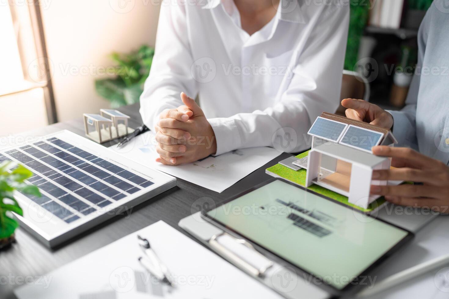 two people sitting at a table with a laptop and a house model solar panel. photo