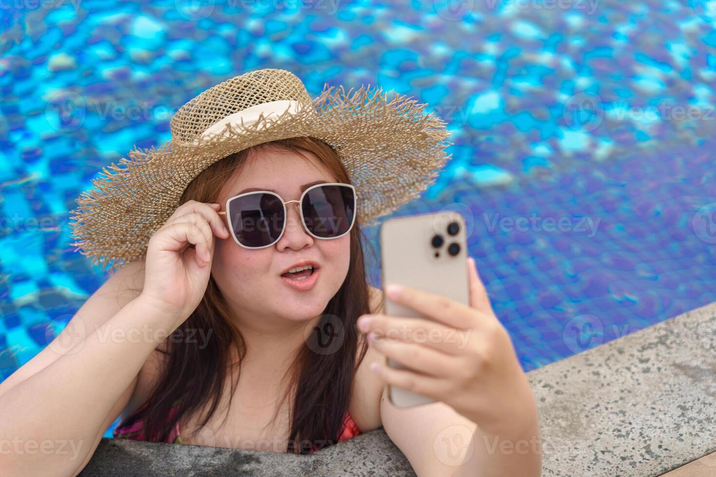 Overweight young woman in a hat and sunglasses taking a selfie in the pool photo