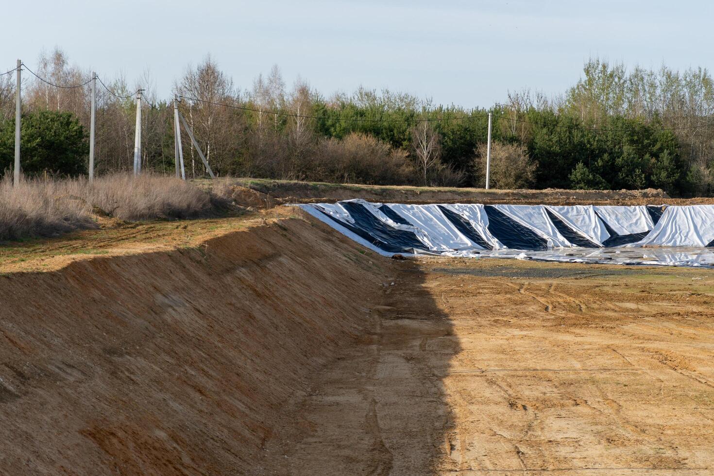 Earthen Pit with Protective Covering, Surrounded by Rural Landscape and Overhead Utility Poles on a Sunny Day photo
