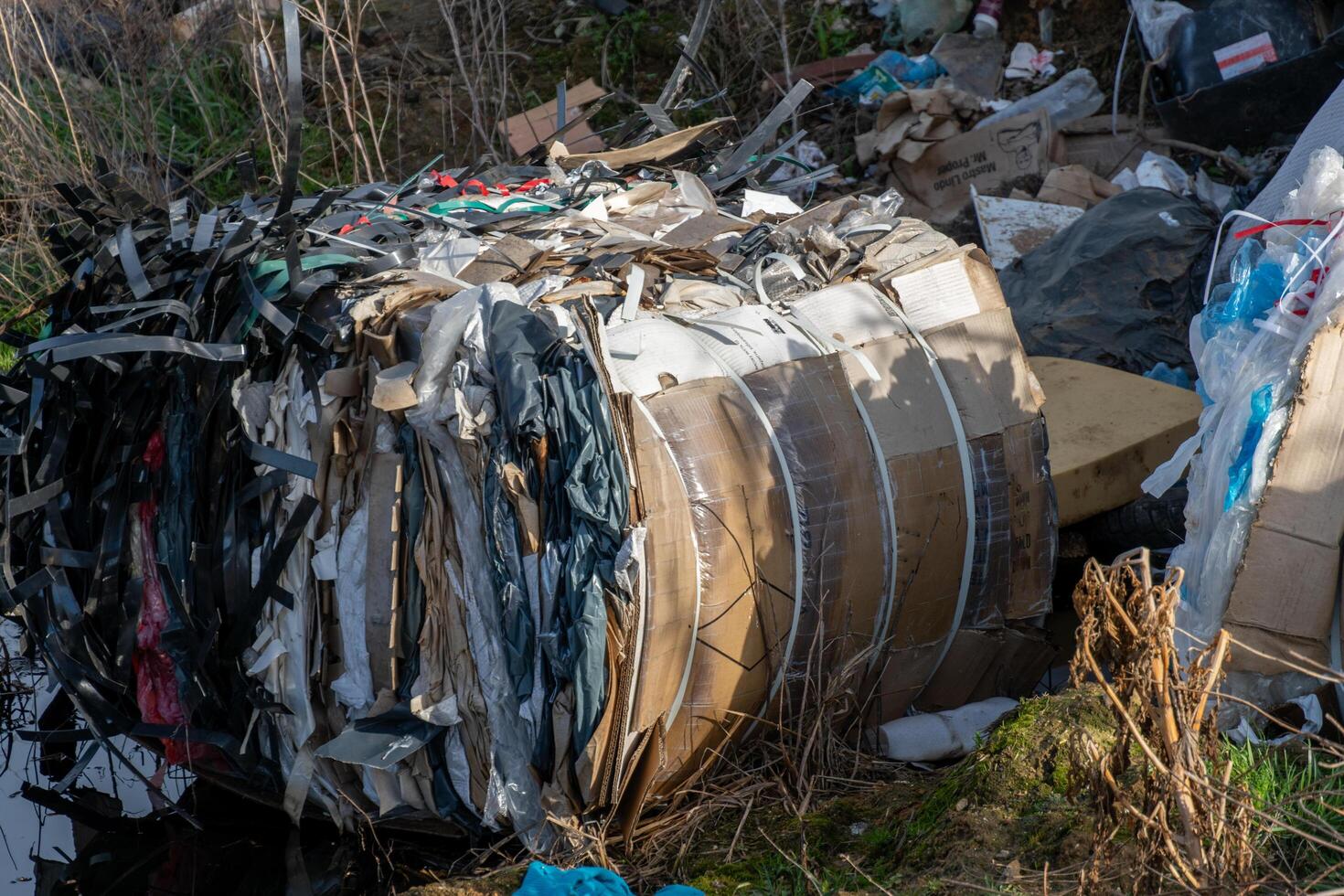 Overstuffed bale of compressed cardboard and plastic packaging materials sits amidst general refuse in an outdoor setting photo