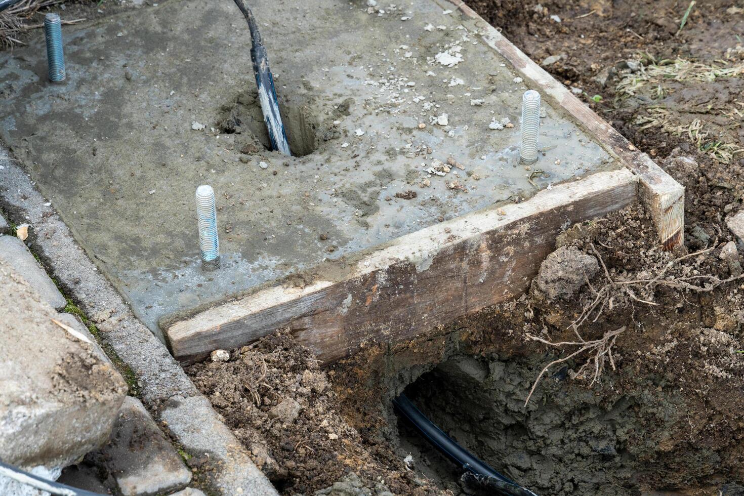 Fresh concrete slab with anchor bolts set in a wooden form during construction, with ground and underground wire conduit visible photo
