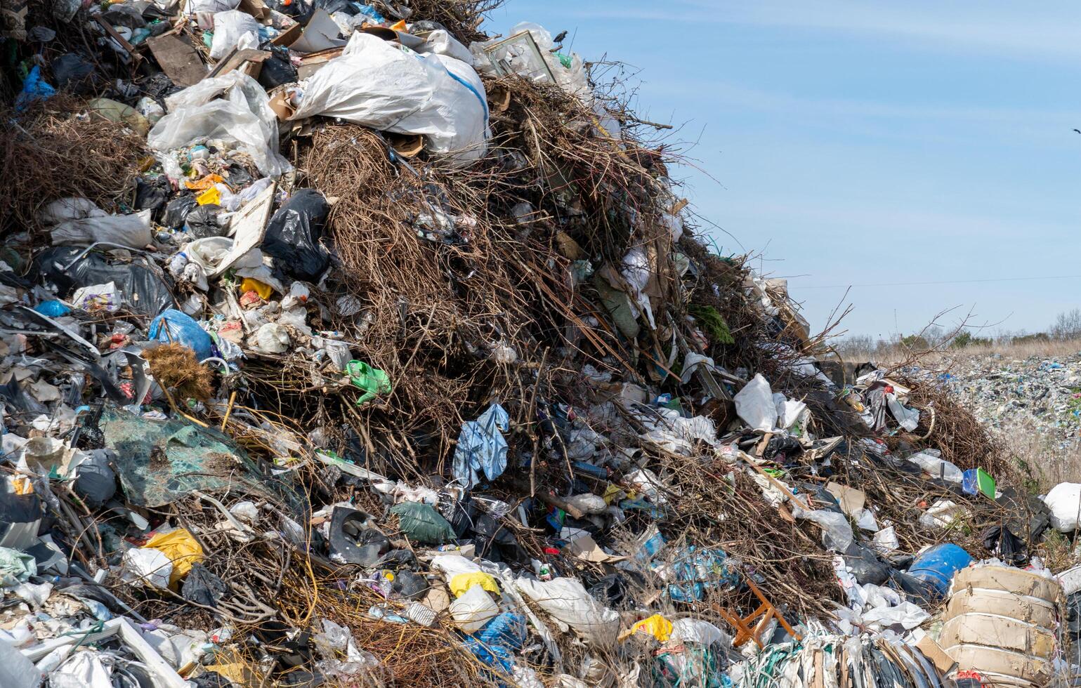 Landfill accumulation showcases extensive mix of discarded trash, vegetation, and plastic waste under clear blue sky photo