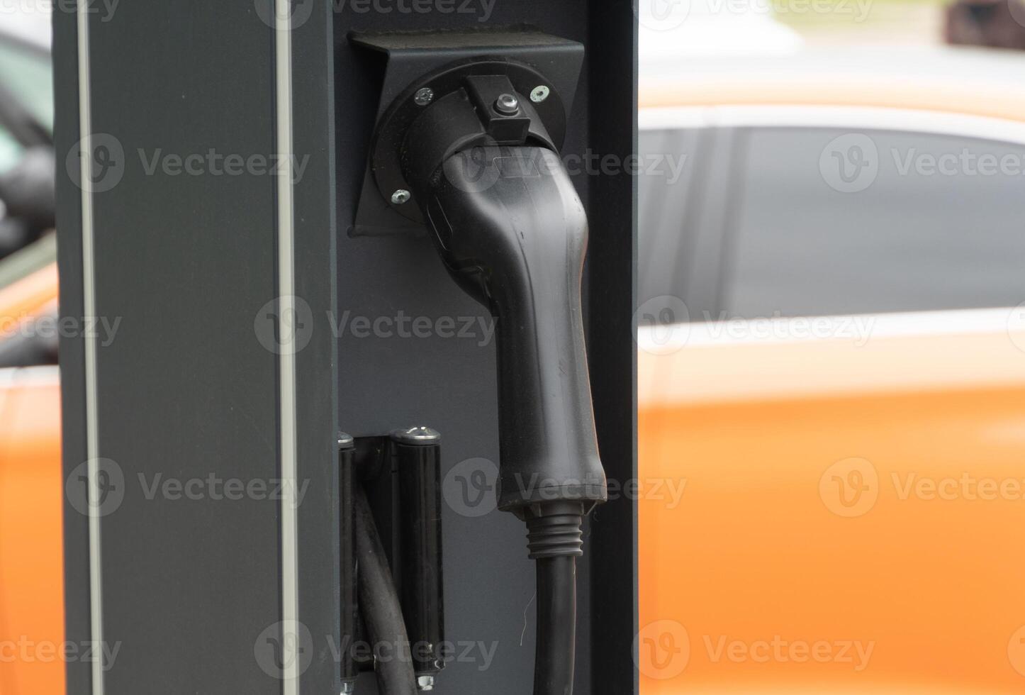 A close-up view of an electric vehicle charger plugged into a charging station in a city environment. The background shows an orange car parked nearby photo