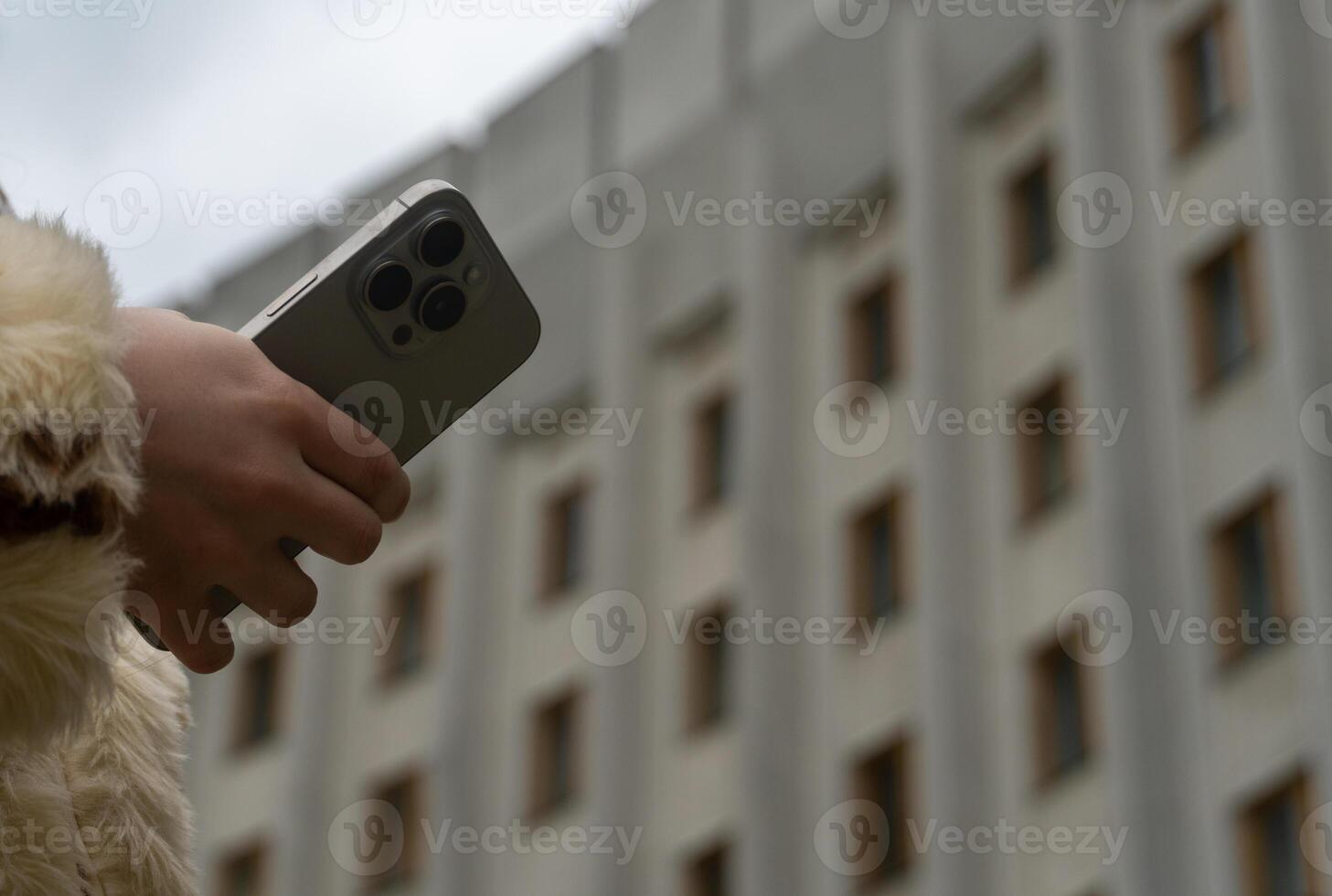 A person with a furry coat holds a smartphone in one hand, standing in front of a contemporary building under a cloudy sky, showcasing urban life photo